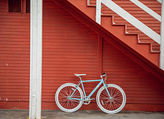 Ein Fahrrad lehnt an der Fassade eines Holzhauses. Die Farben Weiß und Orange dominieren das Bild