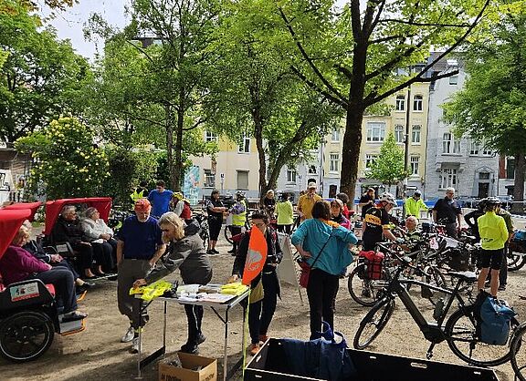 Das Foto zeigt einen Infostand mit vielen Radfahrenden im Schatten unter Bäumen