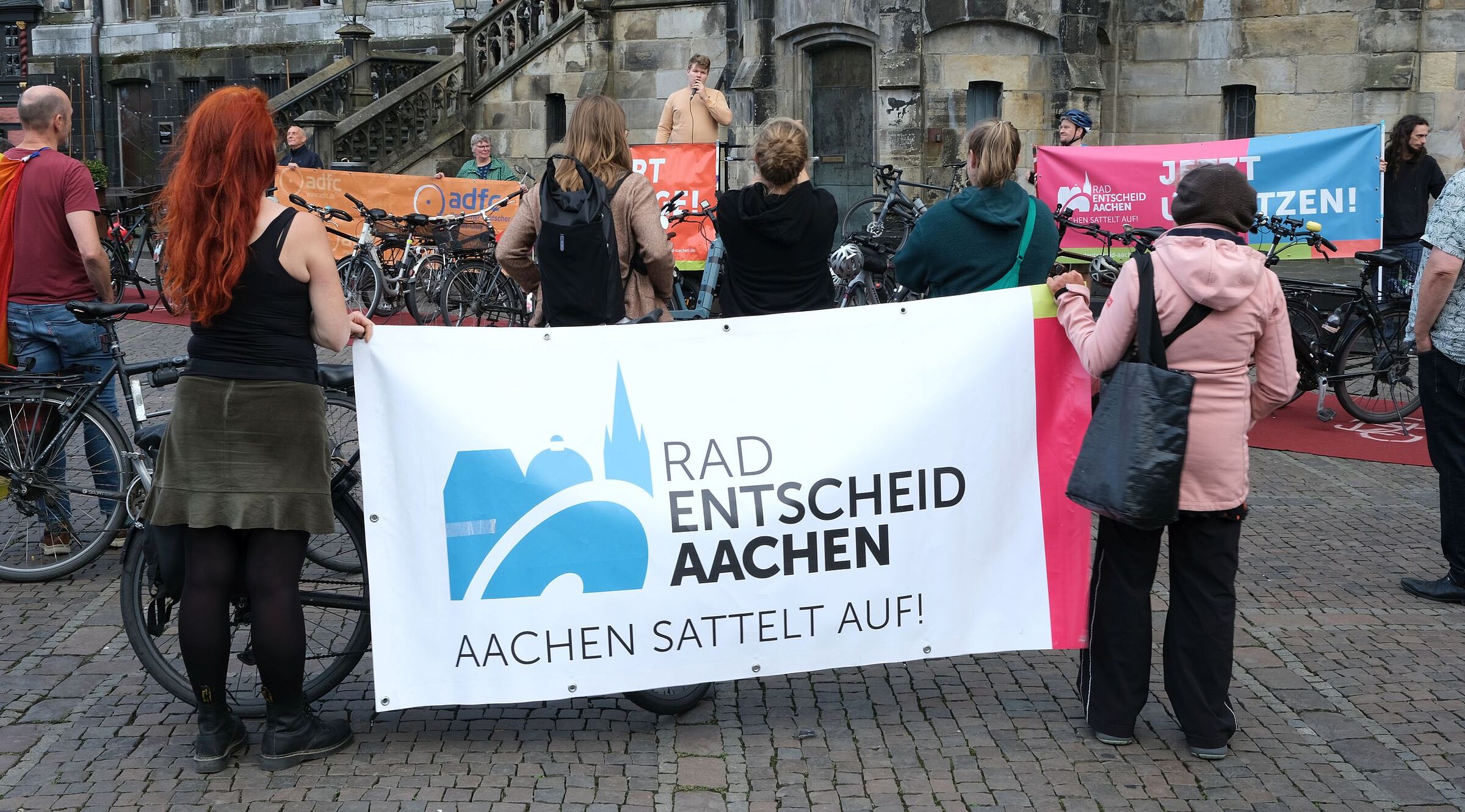 Teilnehmende einer Mahnwache vor dem Aachener Rathaus stehen mit Fahrrädern und Bannern von Radentscheid Aachen und ADFC vor einem Redner mit Mikrofon.