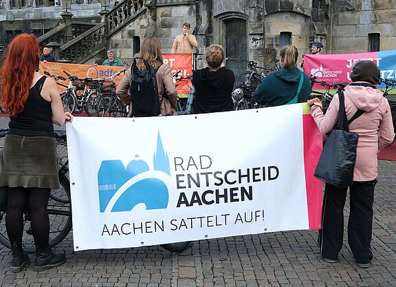 Teilnehmende einer Mahnwache vor dem Aachener Rathaus stehen mit Fahrrädern und Bannern von Radentscheid Aachen und ADFC vor einem Redner mit Mikrofon.