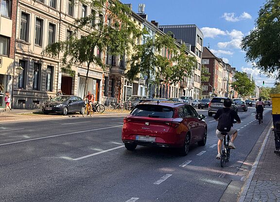 Zwei Radfahrende fahren auf der Wilhelmstraße neben Autos und einem Lieferfahrzeug. Die Radverkehrsführung ist schmal markiert und liegt unmittelbar neben dem Kfz-Verkehr.