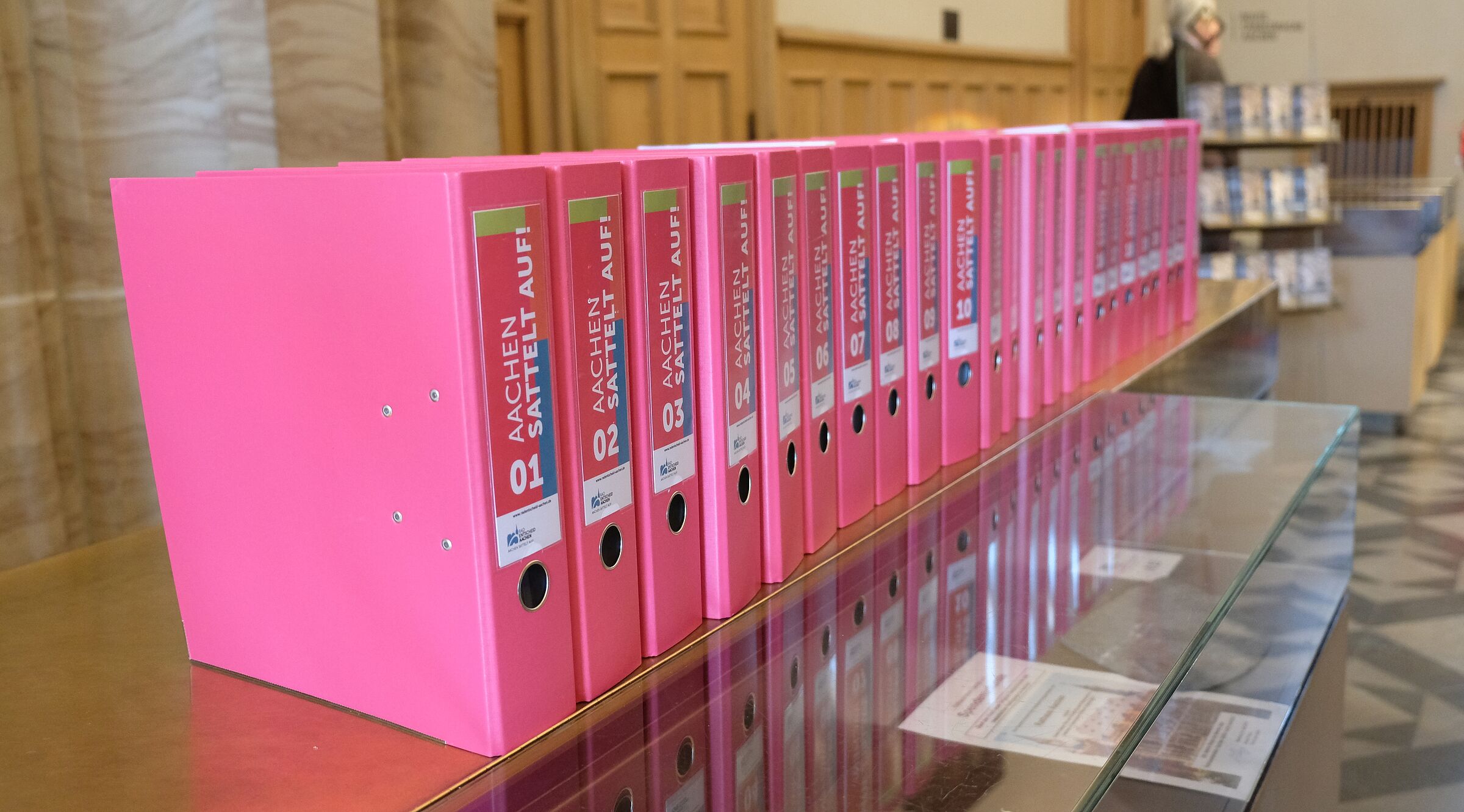 Eine lange Reihe pinker Aktenordner mit der Aufschrift „Aachen sattelt auf“ steht auf einer Glasvitrine im Rathaus. Die Ordner enthalten die gesammelten Unterschriften des Radentscheids Aachen.