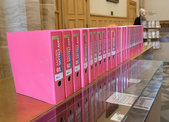 Eine lange Reihe pinker Aktenordner mit der Aufschrift „Aachen sattelt auf“ steht auf einer Glasvitrine im Rathaus. Die Ordner enthalten die gesammelten Unterschriften des Radentscheids Aachen.