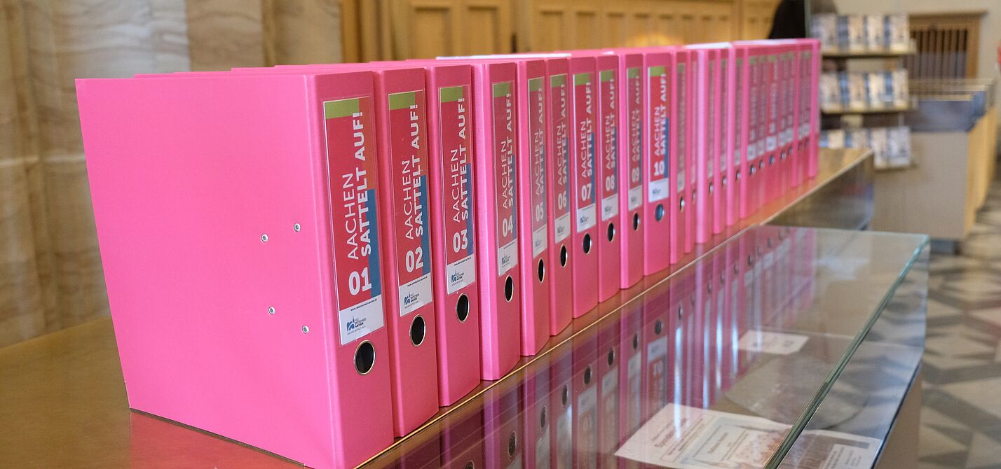 Eine lange Reihe pinker Aktenordner mit der Aufschrift „Aachen sattelt auf“ steht auf einer Glasvitrine im Rathaus. Die Ordner enthalten die gesammelten Unterschriften des Radentscheids Aachen.