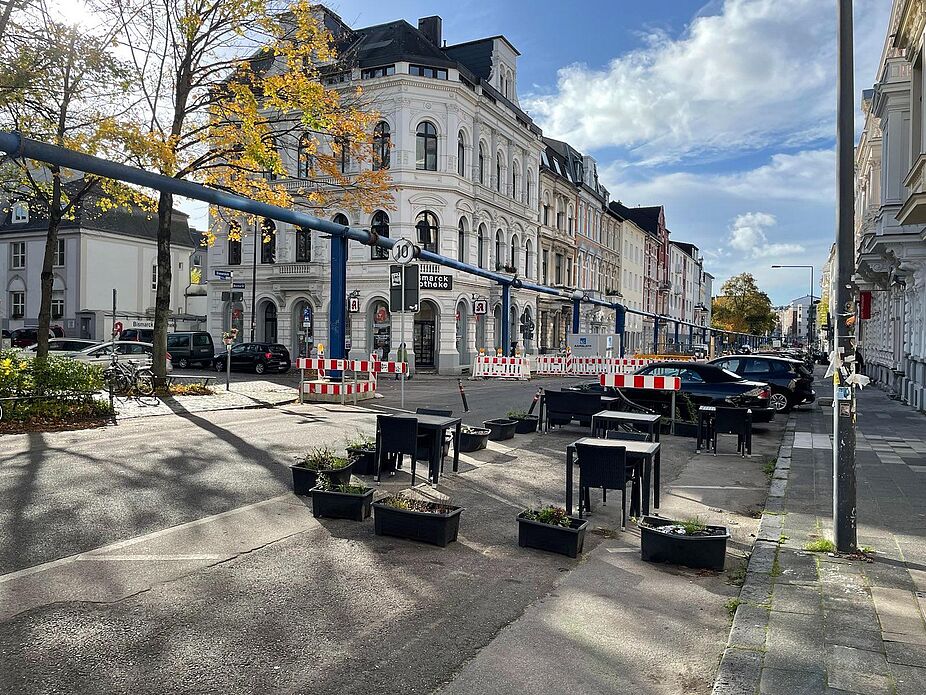 Bismarckstraße am Neumarkt Die Bismarckstraße auf Höhe des Modalfilters am Neumarkt, zu sehen sind auch Sitzmöbel für Außengastronomie. Zudem befinden sich im Hintergrund Warnbaken und Poller