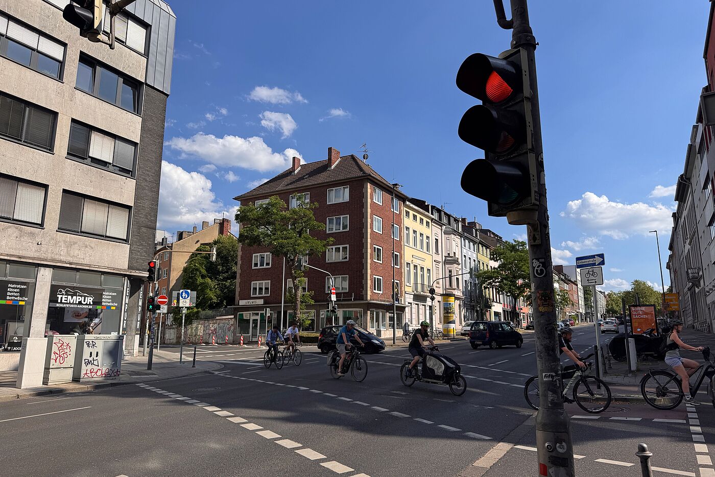 Mehrere Menschen auf Fahrrädern und einem Lastenrad queren eine Kreuzung an der Wilhelmstraße. Die Fahrbahn ist breit, Ampeln und Kfz-Verkehr prägen die Situation.