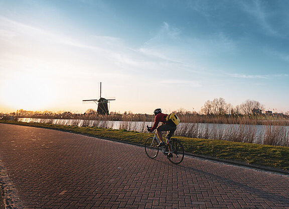 Eine Person fährt in ländlicher Gegend neben einem Fluss auf einem Rennrad der tief stehenden Sonne entgegen. Im Hintergrund ist eine Windmühle zu sehen