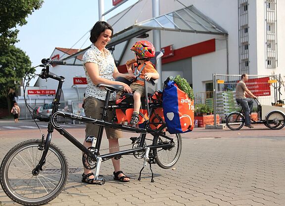 Frau schnallt Kind im Kindersitz an. Es sitzt hinten auf dem Fahrrad, dahinter sind noch Gepäcktaschen angehängt.