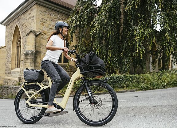 Fröhliche Frau fährt auf Elektrofahrrad, im Hintergrund eine Kirche. 