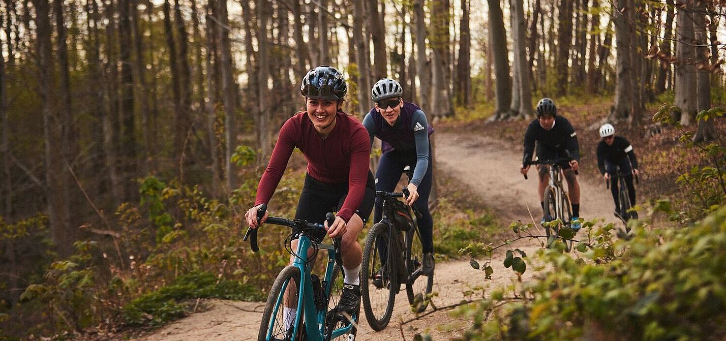 Eine Gruppe von Frauen und Männern fahren mit Gravelbikes hintereinander auf einem Waldweg. 